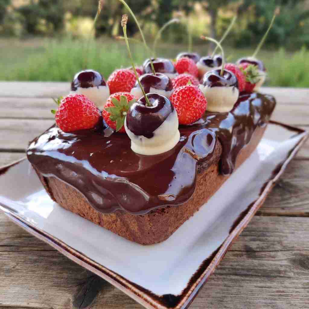 image of a cake loaf covered in a dark chocolate glaze and topped with fresh strawberries and cherries dipped in white chocolate, you can find the recipe on the blog og Gourmandises Academie
