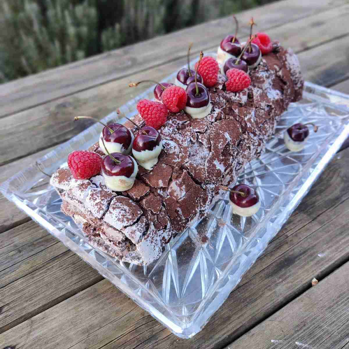 picutre of a meringue roulade on a glass plate, dusted with cocoa and decorated with raspberries and cherries dipped in dark chocolate, the recipe can be found on the blog of cookery school gourmandises academie, cambridge