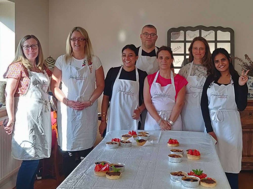 A group of 7 people standing behind a table, showcasing their tartelettes, during a team bonding session at the cookery school Gourmandises Academie, Cambridge
