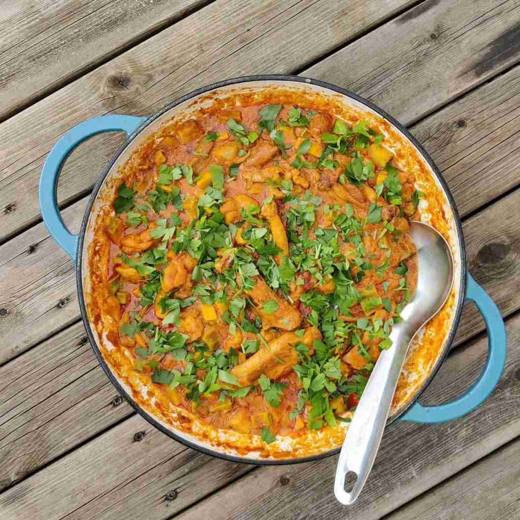 image with a blue pan on a wooden table, filled with bits of chicken in a tomato sauce and parsley on top, recipe available at cookery school Gourmandises Academie, Cambridge