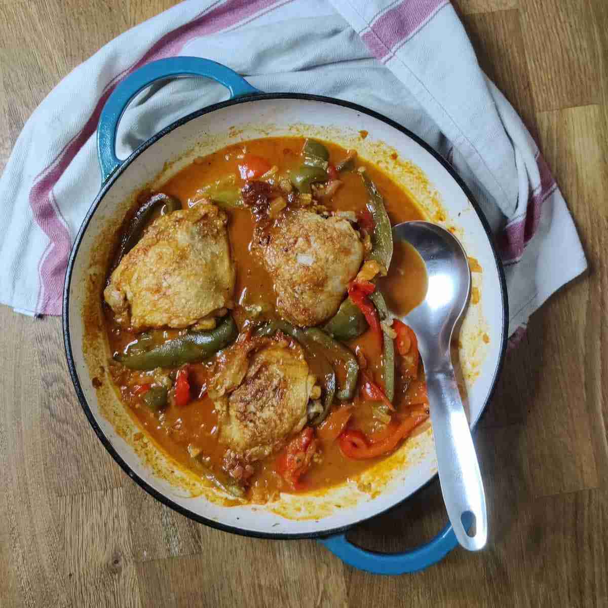 picture of a blue casserole dish on top of a towel with red stripes, with pieces of chicken, red and green peppers in a tomato sauce, the recipe is available on the blog of the cookery school Gourmandises Academie, Cambridge