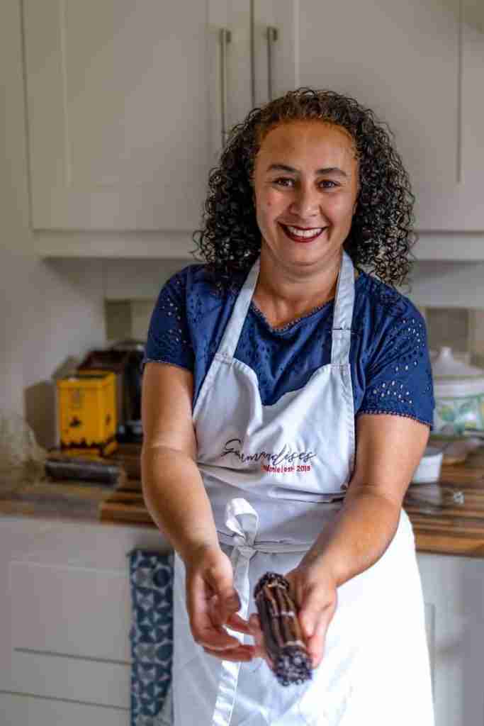 Image of a lady, in a kitchen, with an apron, smiling and holding a bunch of vanilla pods, at cooking school Gourmandises Academie