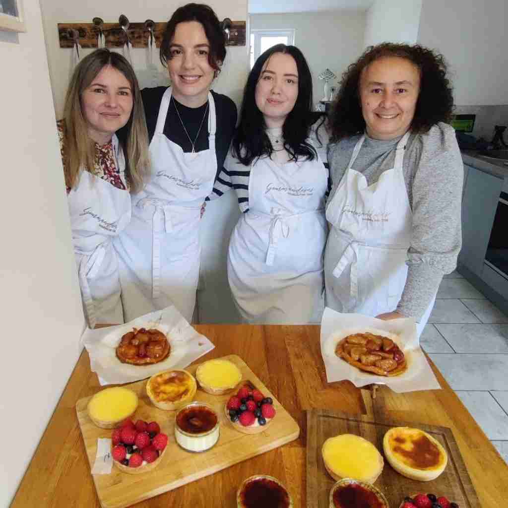 image of fours ladies smiling in front of an array of desserts, tarte au citron, flan patissier, tarte tatin, creme brulee and tarte au fruits made during a baking class at gourmandises academie, cambridge